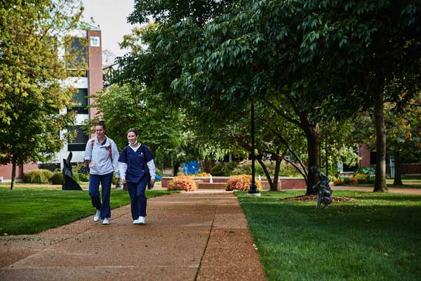 Two students in walking on a sidewalk in front of trees and campus buildings