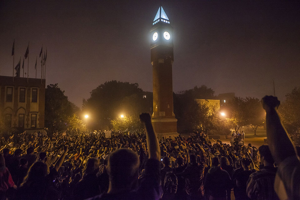 The SLU Clock Tower is lit up at night while surrounded by students with fists raised in the air.