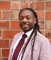 Richard Marks, wearing a pink suit, maroon tie and white shirt, stands in front of a brick wall and smiles