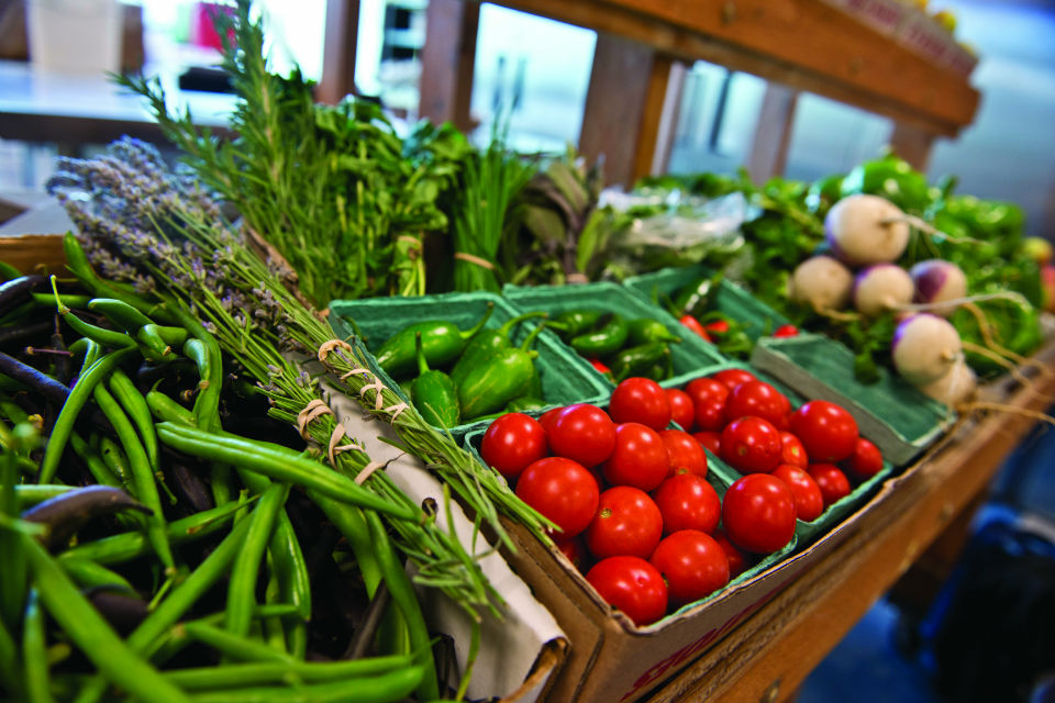 A row of fresh produce in baskets at a market including peppers, tomatoes, onions and greens