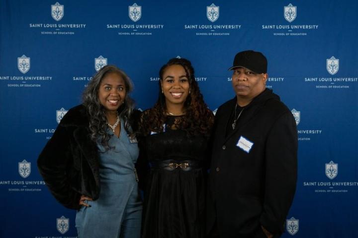 Thompson Three people smile at the camera while standing in front of a Saint Louis University backdrop