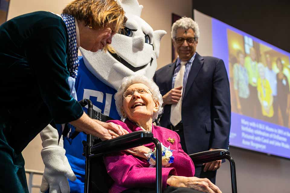 Two people stand and one person sits on a stage near the Billiken.