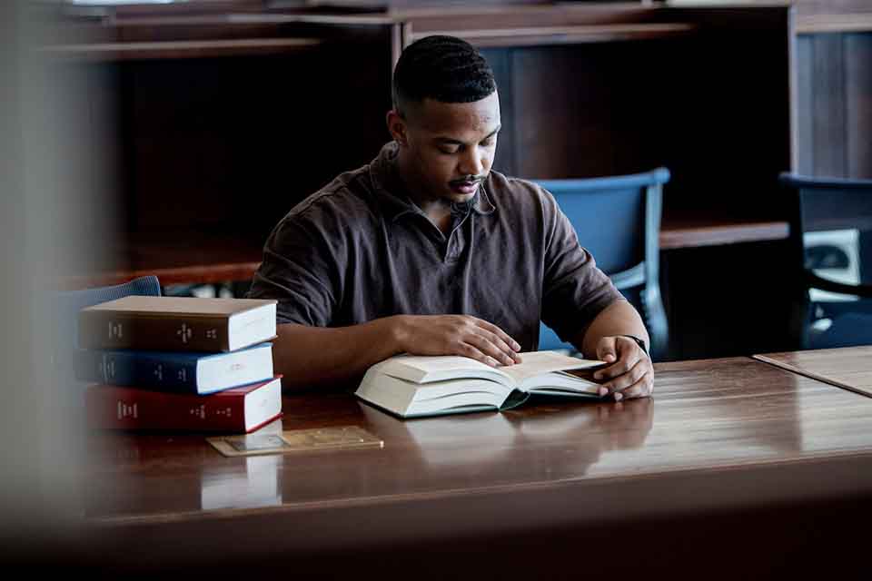 A male student reads from a book at a table in thel library.