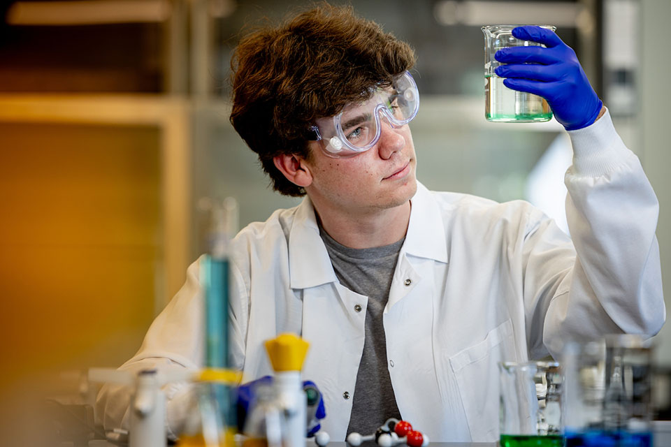 A student wearing a lab coat and googles looks closely at a glass container filled with liquid in a lab.