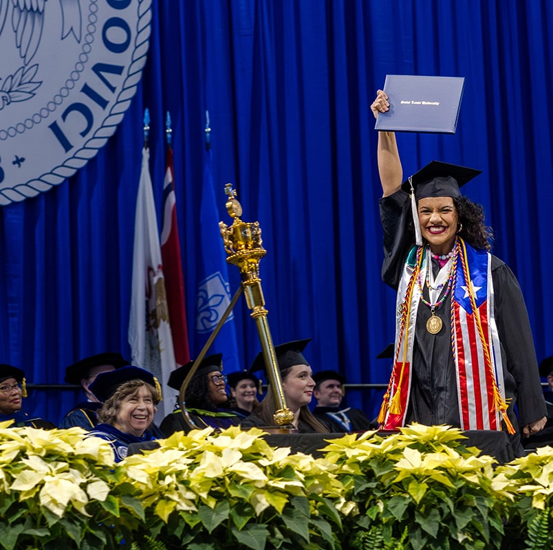 a student wearing graduation cap and gown holds her diploma in the air with a huge smile on her face.
