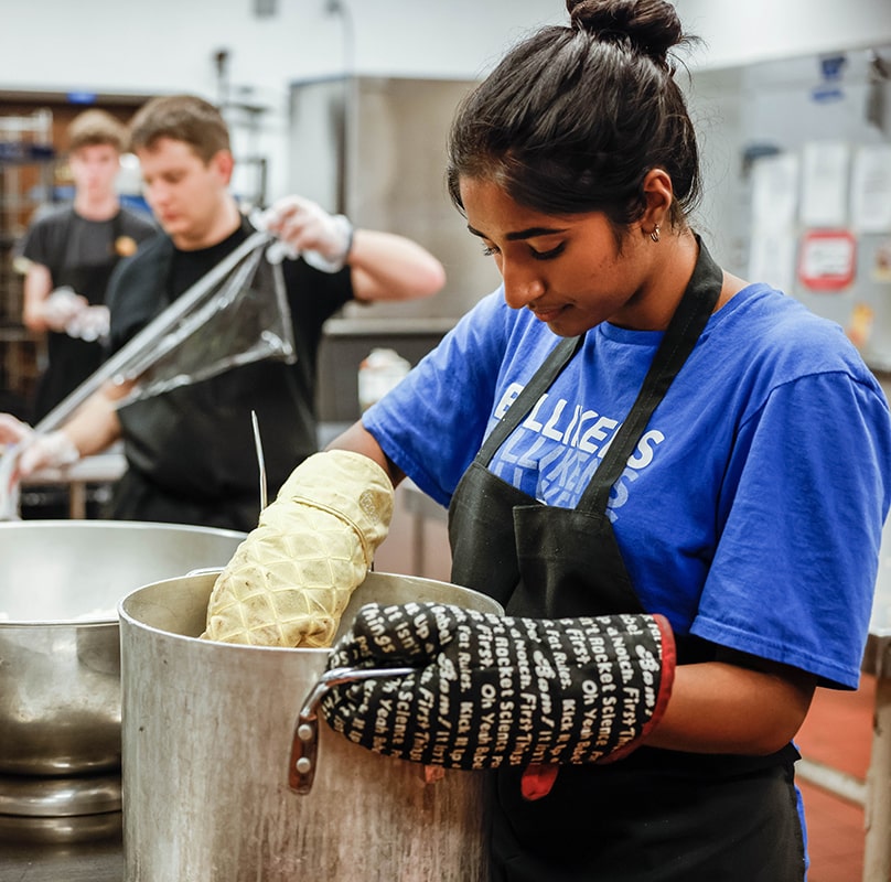 A student wearing an apron and oven mitts works at a large metal pot while standing in a commercial kitchen. Another student behind her unrolls saran wrap.