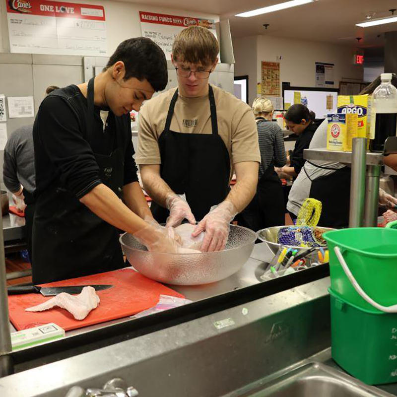 Two students work with a turkey in a bowl in a commercial kitchen.
