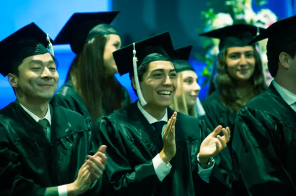 A student, dressed in a graduation gown, shakes hands with a SLU faculty members on stage during commencement. American and Spanish flags are in the background.