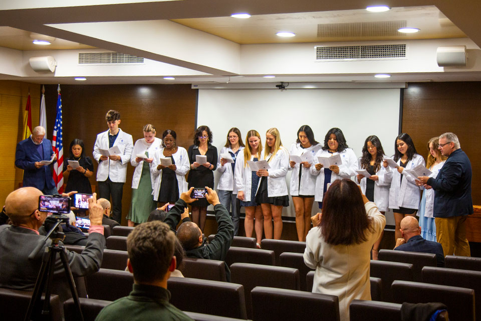 SLU-Madrid Celebrates 9th Annual White Coat Ceremony Students wearing white coats and faculty members read from pieces of paper at the front of an auditorium.