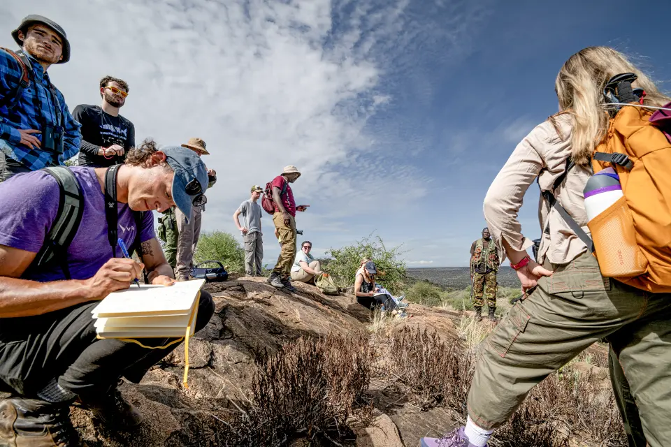 Man standing on top of mount pointing to surrounding areas while participants take notes in notes or listen and look toward him. 