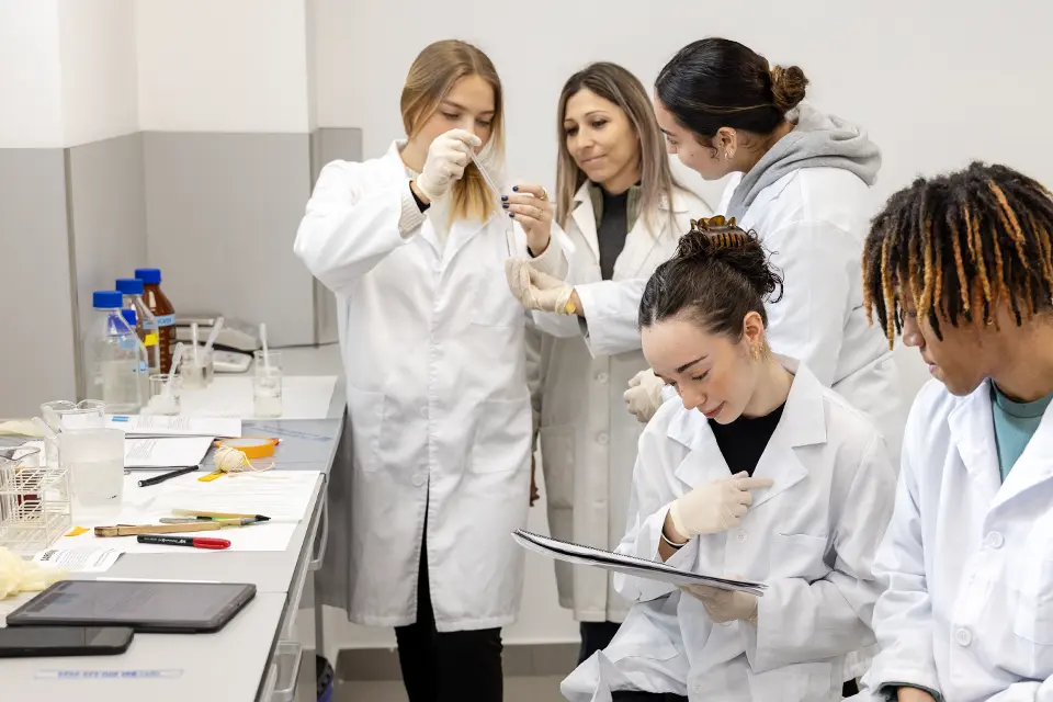 Student holding specimen speaks with professor and fellow student while two other students look at notebook in a science lab.