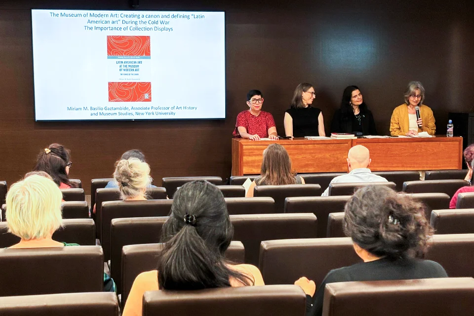 Fabiola Martínez, Ph.D. gives opening remarks sitting next to (from right to left): Laura Katzman, Ph.D., Beatriz Cordero Martín, Ph.D. and keynote speaker Miriam Basilio Gaztambide, Ph.D. Four panelists sit at the front of a lecture hall while one speaks into a microphone. A screen displays a slide with the title: The Museum of Modern Art: Creating a Canon and Defining 'Latin American Art' During the Cold War — The Importance of Collection Displays