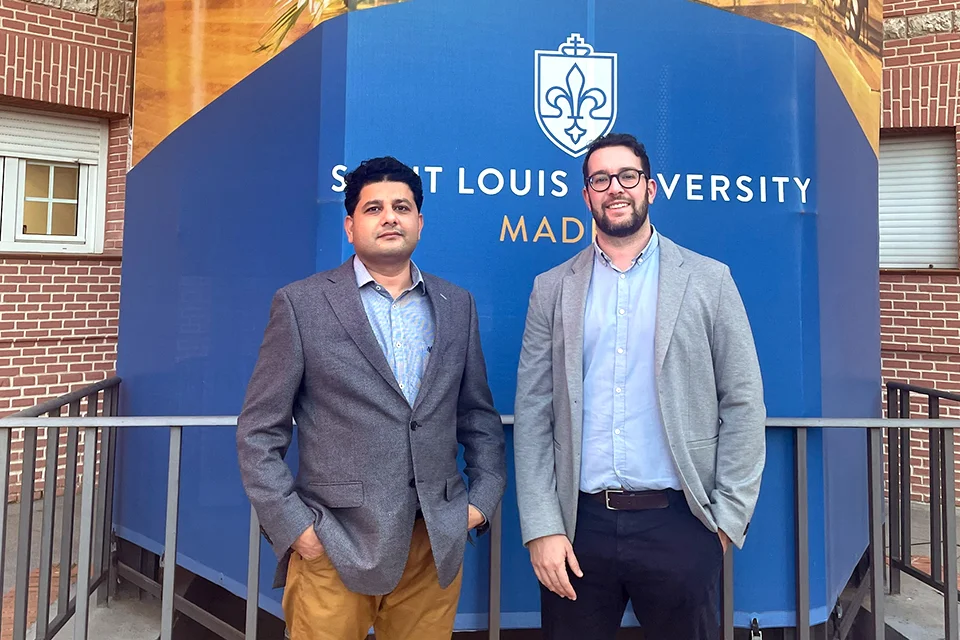 Professor Rehman and PUE's Àngel Garcia pose outside the stairwell in Padre Arrupe Hall patio with the University logo behind them on the banner wrapping around the stairs. 