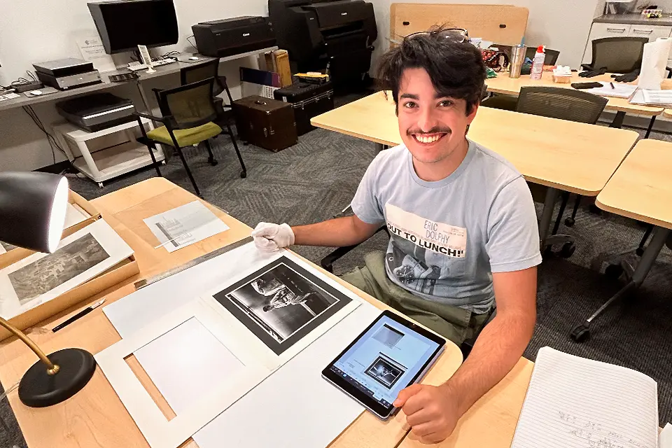 Student posing in his office with a laptop and photography tools on the table.