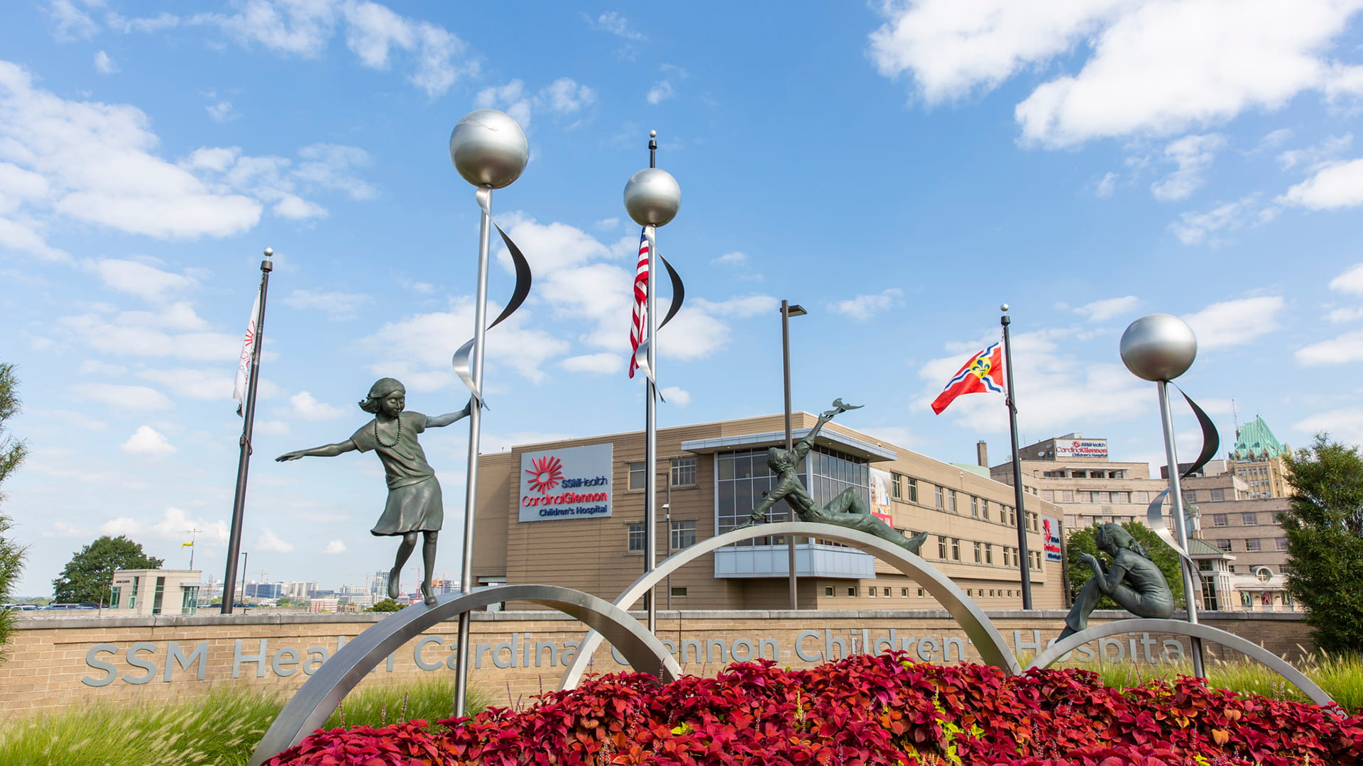 Exterior image of Cardinal Glennon Children's Hospital with red flowers in the foreground, flag poles and statues of playing children in front of the building.