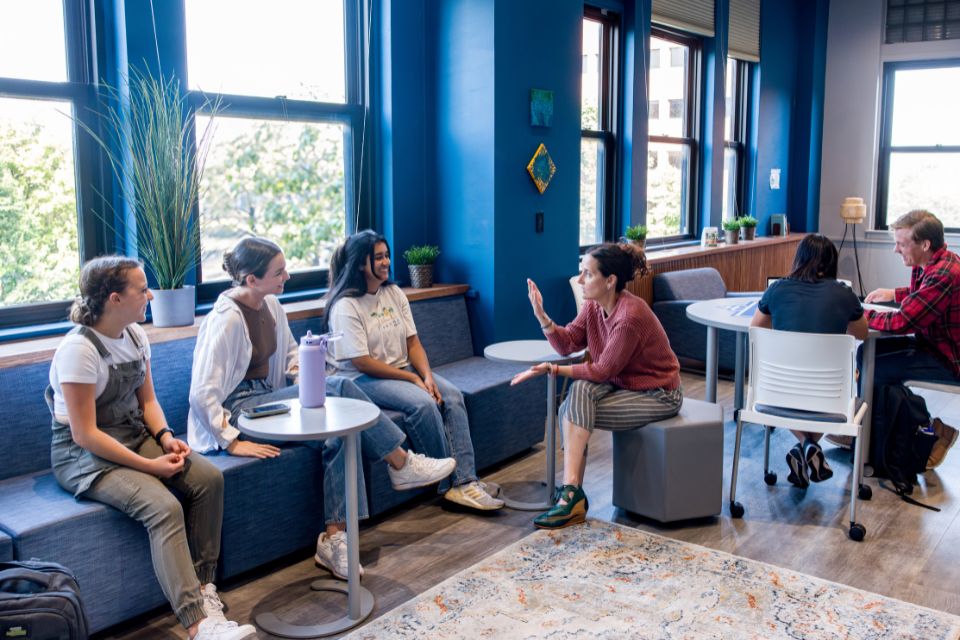 Students and a faculty member sit on chairs and benches in a room with windows and plants.