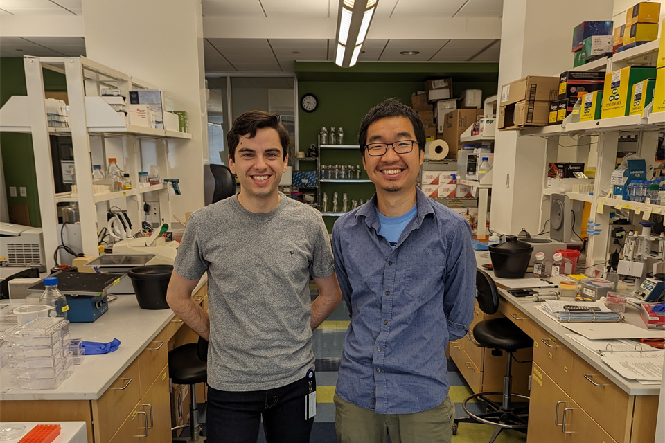 Two researchers stand in the middle of a lab, surrounded by equipment and supplies.