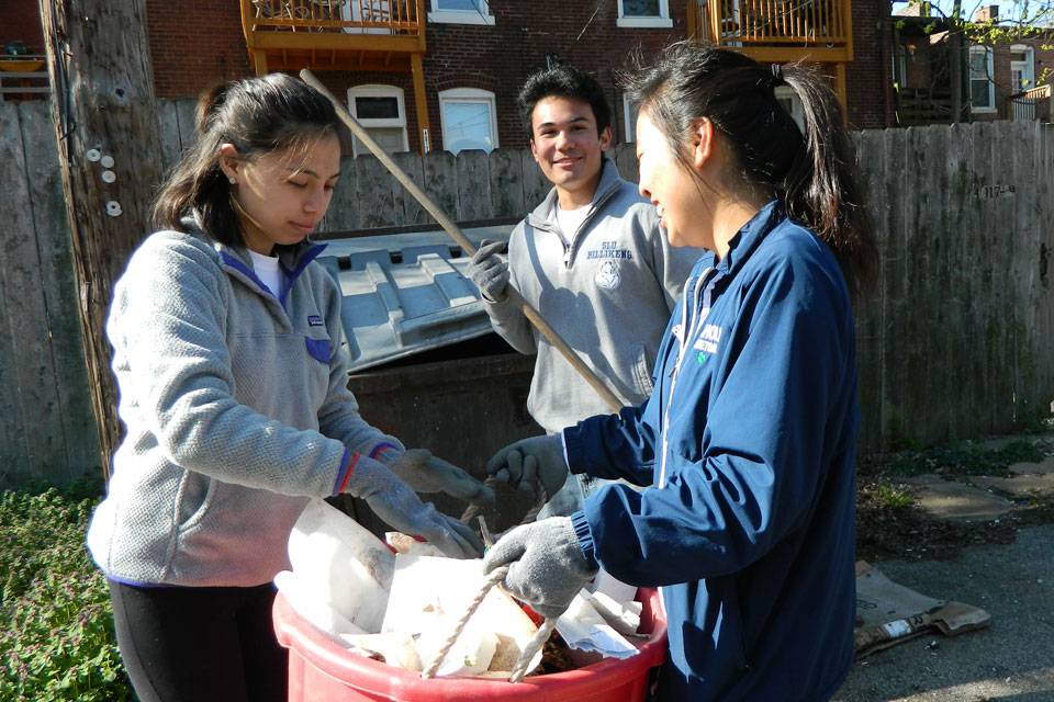 Two students wearing work gloves examine the contents of a bucket while another student holds a work tool in the background.