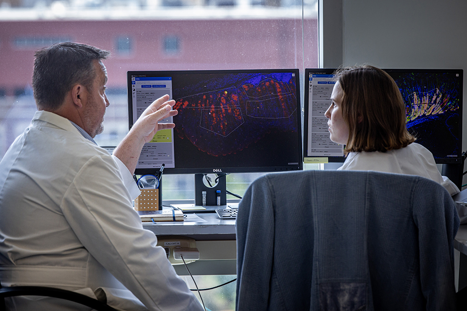 A photo of two people inside a lab wearing white coats. They sit in front of computer monitors. They discuss colorful data displayed on the screen.  