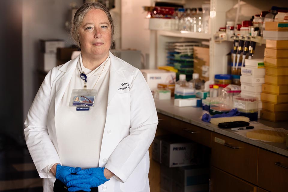 Dr. Sarah George poses for a photo inside a lab. She is wearing a white shirt under a white lab coat. A badge is clipped to her collar. Her hands are folded at her waist. She is wearing blue gloves. Her long hair is tied back.