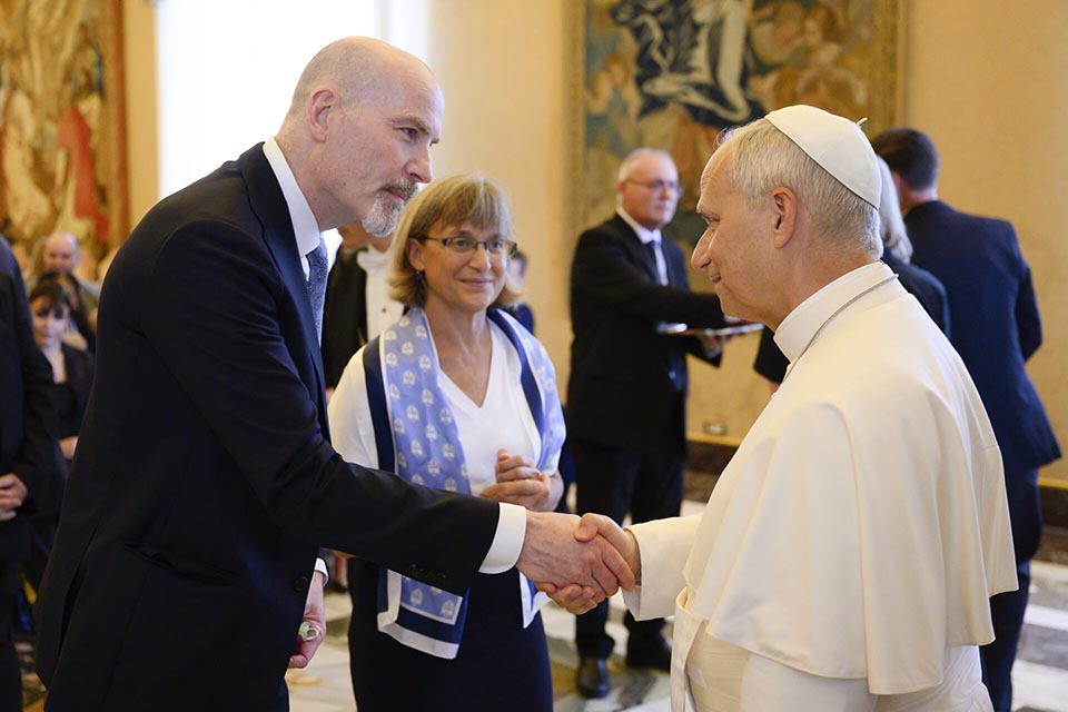 In an ornate room at the Apostolic Palace in Rome, SLU President Edward Feser, wearing a dark suit, shakes hands with Pope Leo XIV, wearing a white papal floor-length robe and cap, as Kathy Feaser looks on, wearing a white top and blue scarf. In an ornate room at the Apostolic Palace in Rome, SLU President Edward Feser, wearing a dark suit, shakes hands with Pope Leo XIV, wearing a white papal floor-length robe and cap, as Kathy Feaser looks on, wearing a white top and blue scarf.