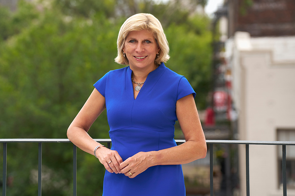 Dr. Kathy Johnson, with blond hair and wearing a royal blue dress, stands confidently on a balcony, smiling. Green trees and a blurred building form the background. Dr. Kathy Johnson, with blond hair and wearing a royal blue dress, stands confidently on a balcony, smiling. Green trees and a blurred building form the background.