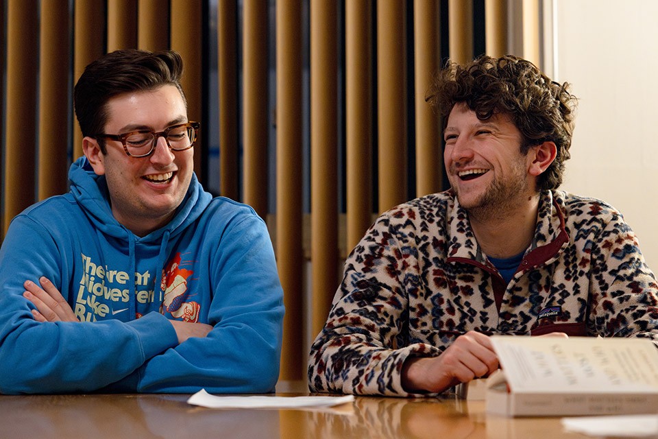 Two students seated at a table smiling and engaged in conversation. One wears a blue hoodie; the other, a patterned fleece. Mood is cheerful and relaxed.