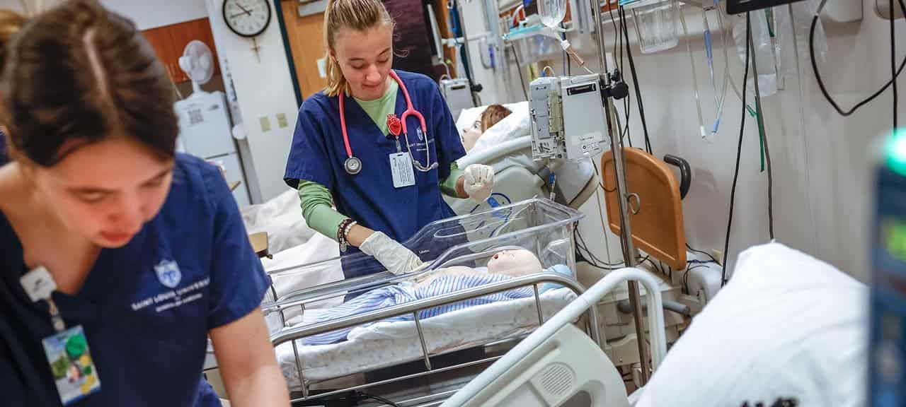 A female student, wearing scrubs and and a stethoscope, examines an infant manikin lying in a hospital bassinet. Another female student, also wearing scrubs, can be seen in the foreground.