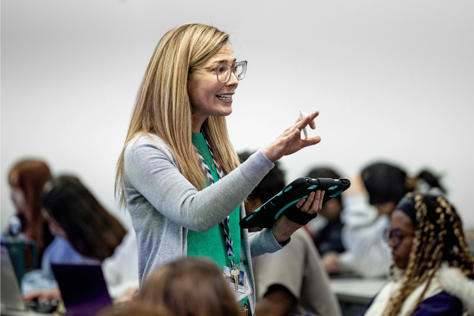 A nursing instructor, wearing a green top and holding an iPad, speaks to a classroom of students. 