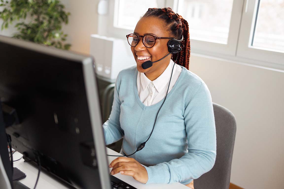 An instructor sits at a desk wearing headphones and smiling while talkig to a computer screen.