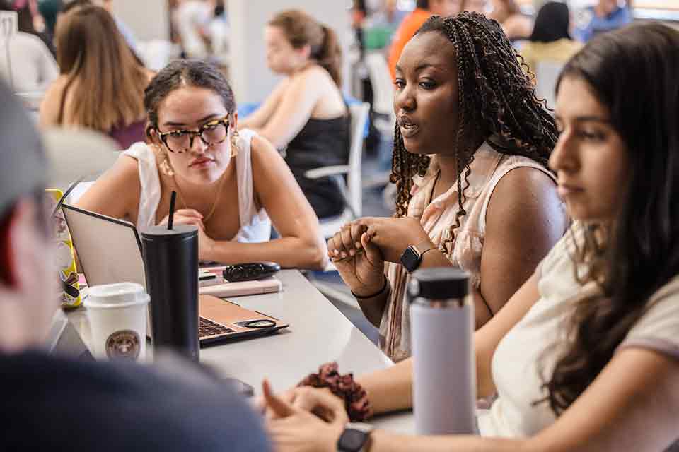 A female student chats with a group of three other students at a desk in a classroom.