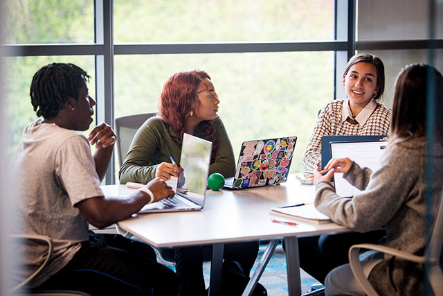 Four students with laptops sit at a table talking. 