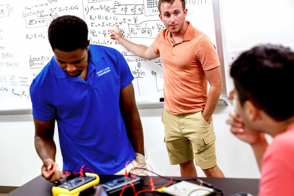 Three students standing in front of a white board working on a physics project