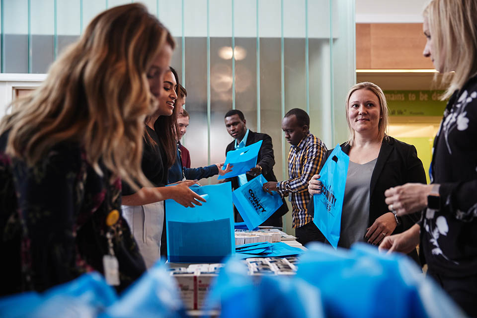 People stand on both sides of a long table handling boxes and blue bags that say Saint Louis County