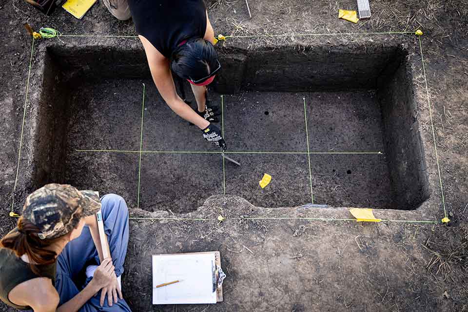 Two students measure out a large, rectangular hole that has been dug into the ground as they prepare to extract an underground artifact at the Cahokia Mounds Historical Site.