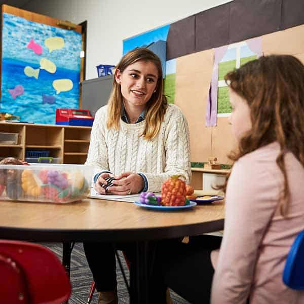 A female student talks with a young girl at a small round table in a classroom